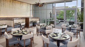 A modern dining area with multiple tables and chairs, large windows offering a view of the outdoors, and a contemporary ceiling light fixture. - Hotel Indigo Flushing - LaGuardia by IHG (Flushing)