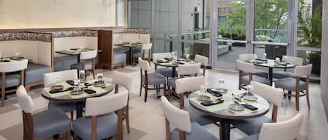 A modern dining area with multiple tables and chairs, large windows offering a view of the outdoors, and a contemporary ceiling light fixture.
