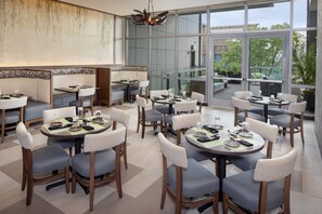 A modern dining area with multiple tables and chairs, large windows offering a view of the outdoors, and a contemporary ceiling light fixture. - Hotel Indigo Flushing - LaGuardia by IHG (Flushing)