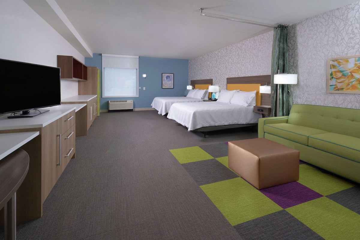 A hotel room with two beds, a desk with a TV, a green sofa, and a small table.