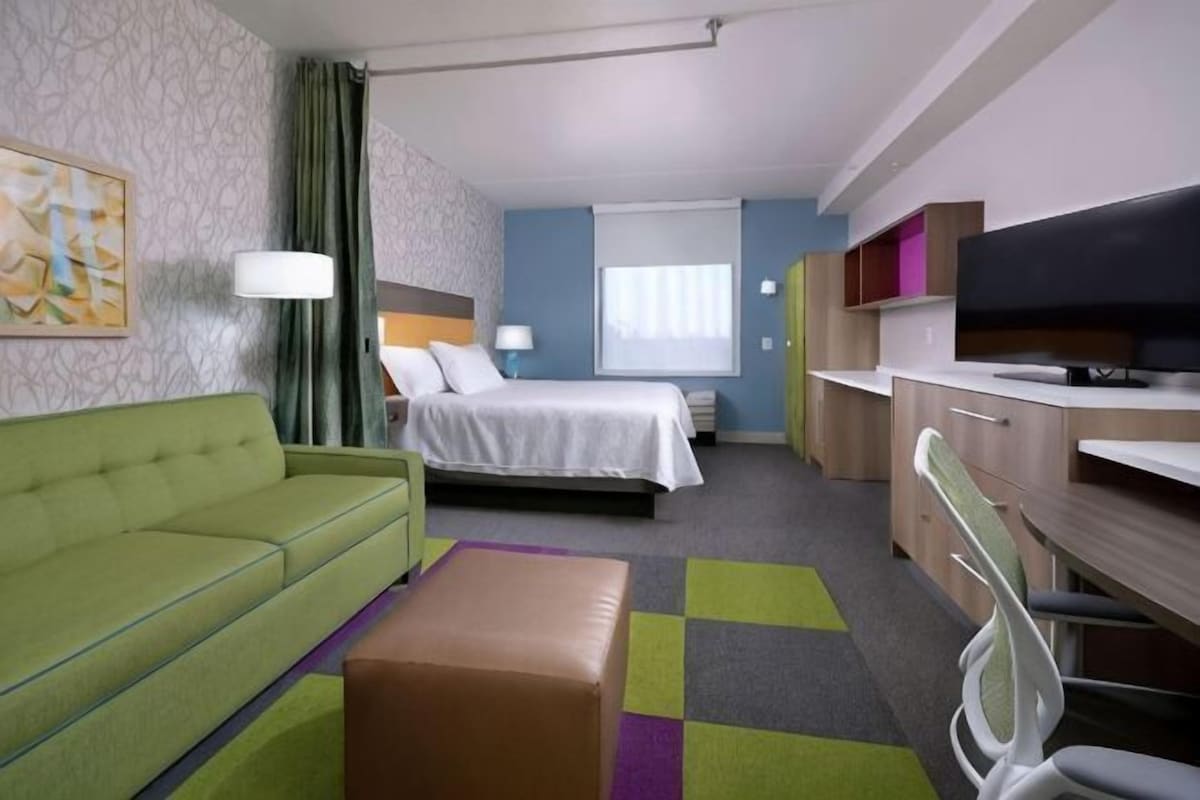 A hotel room with a green sofa, a bed, a desk with a chair, a television, and a window.