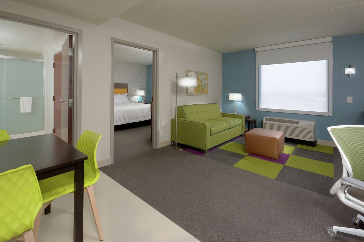 A hotel room with a bed, a green sofa, a small table with chairs, a window with blinds, and an air conditioning unit.