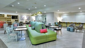 A modern lounge area with a green sofa, multiple tables, chairs, and a flat-screen TV. - Home2 Suites by Hilton Duncan (Duncan)