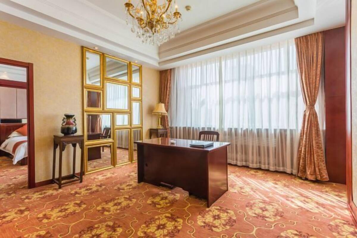 A hotel room with a large desk, a mirror, a chandelier, and a large window with curtains.