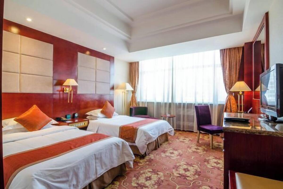 A hotel room with two beds, a TV, a desk, and a window with curtains.