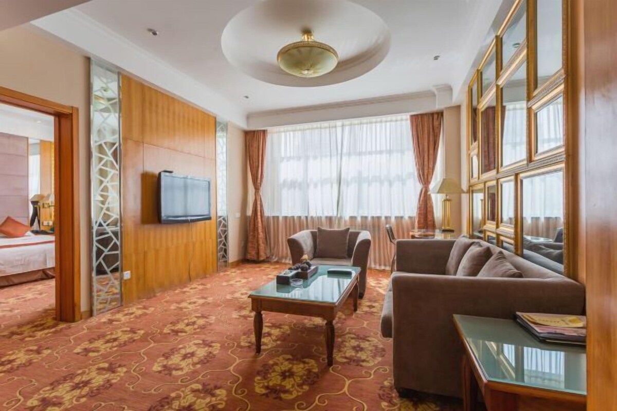 A hotel room with a large bed, a sitting area with a sofa and armchairs, a television, and a large window with curtains.