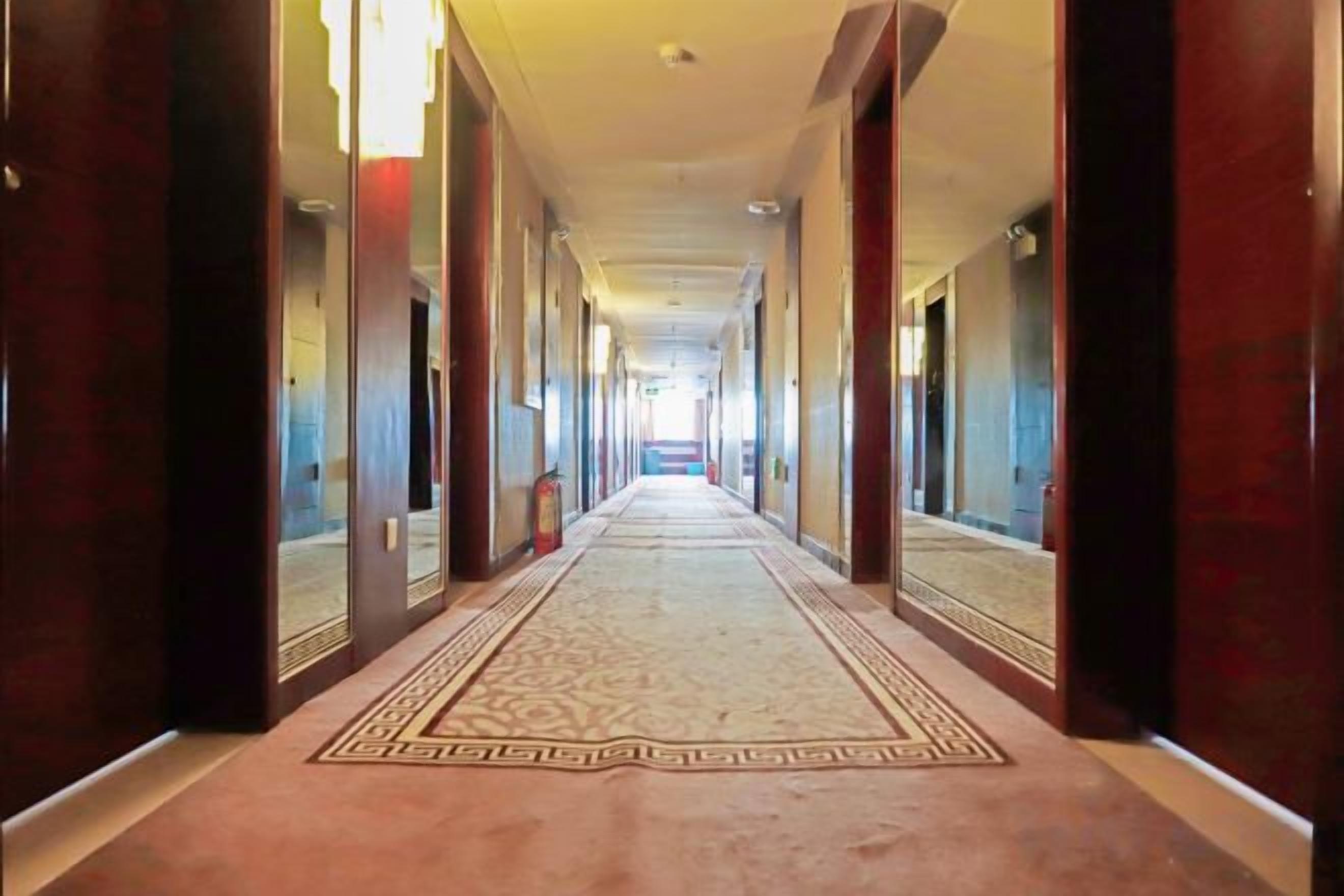 Ein langer, schmaler Hotelkorridor mit Teppichmuster, Holztüren und einer Spiegelwand an einer Seite.