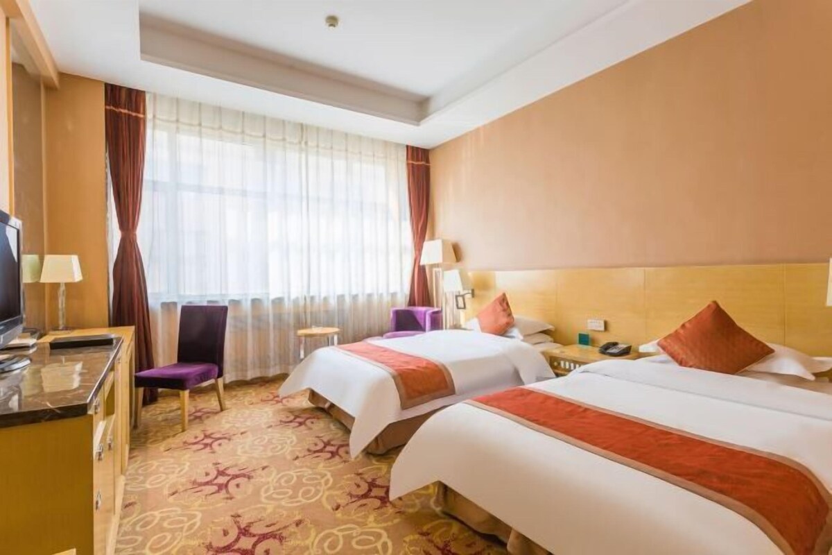 A hotel room with two beds, a desk, a chair, a TV, and a window with curtains.