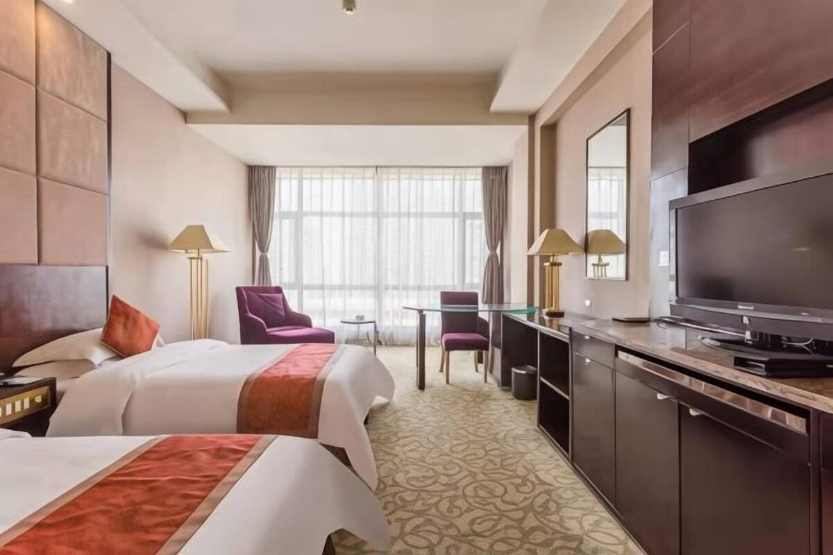 A hotel room with two beds, a desk, a chair, a television, and a large window with curtains.