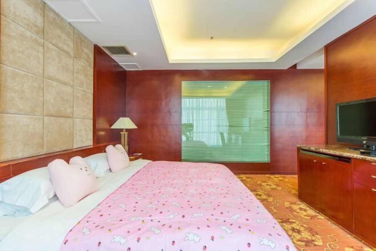 A bedroom with a bed, a television, a window with blinds, and a lamp.