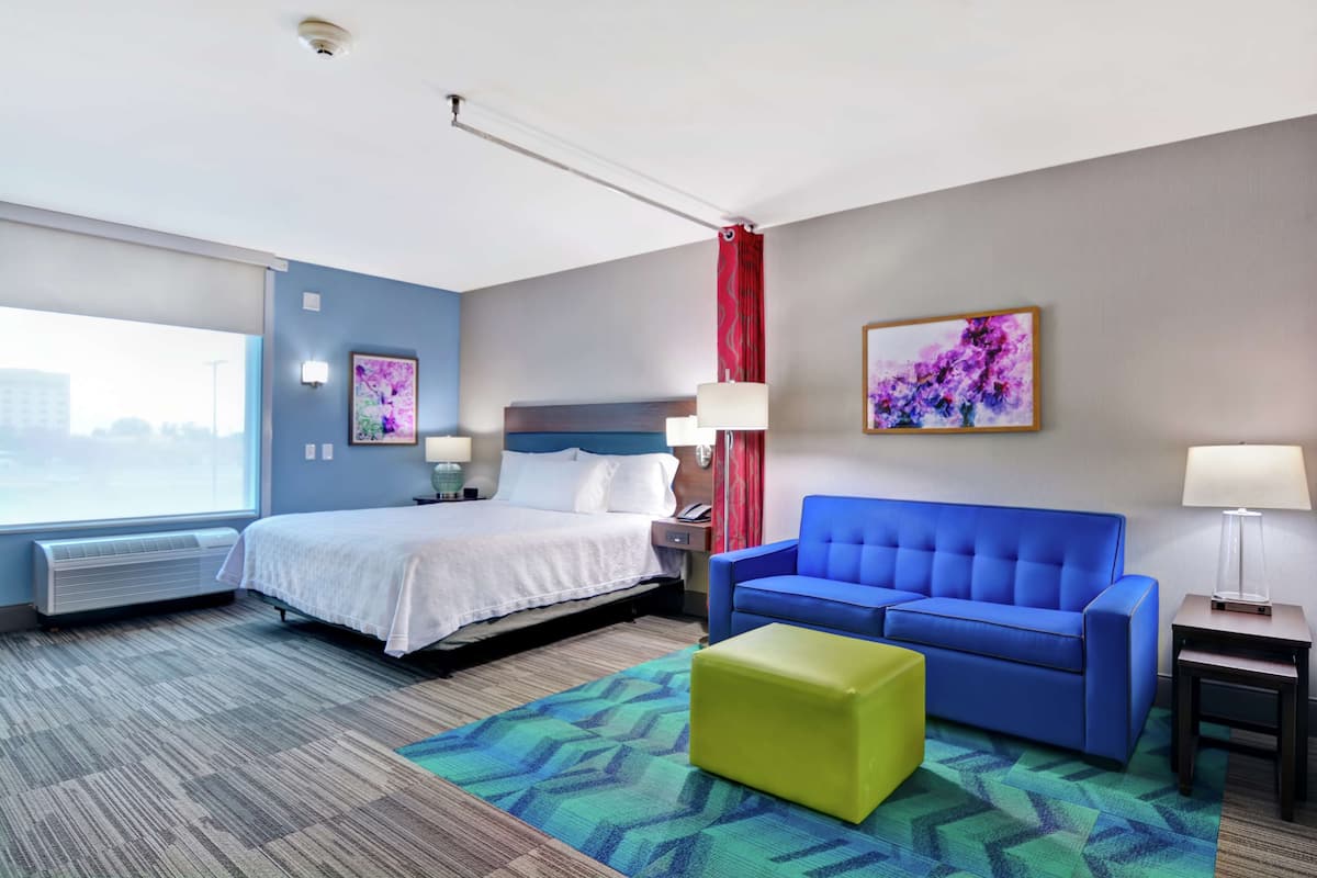 Ein Hotelzimmer mit einem Bett, einem blauen Sofa, einem grünen Hocker, einem Fenster mit Aussicht und einem Bild an der Wand.