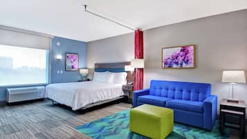 A hotel room with a bed, a blue sofa, a green ottoman, a window with a view, and a painting on the wall.