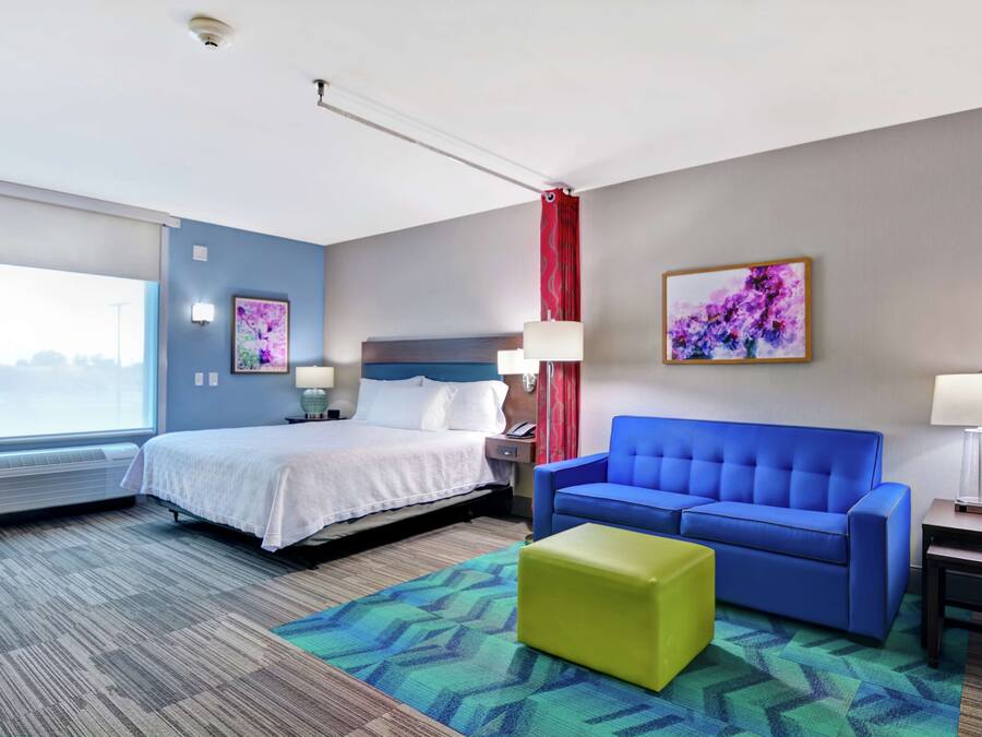 A hotel room with a bed, a blue sofa, a green ottoman, a window with a view, and a painting on the wall.