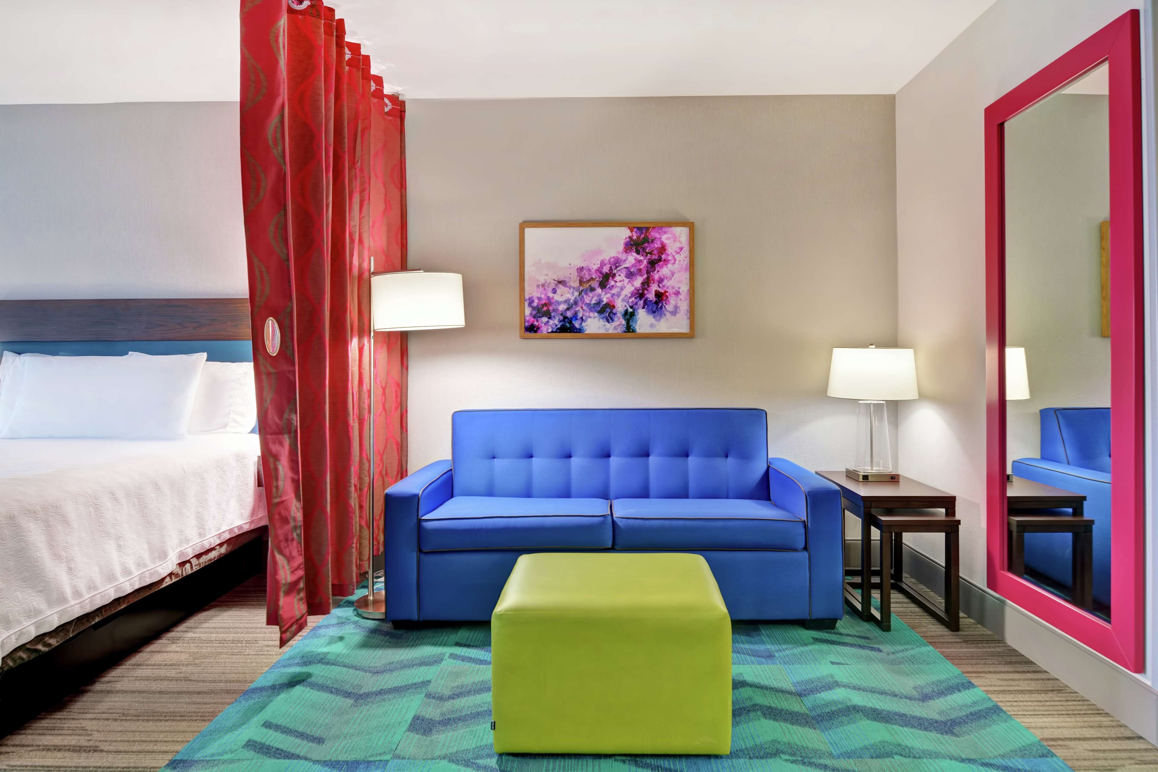 A hotel room with a bed, a blue sofa, a green ottoman, a painting on the wall, and a mirror with a pink frame.