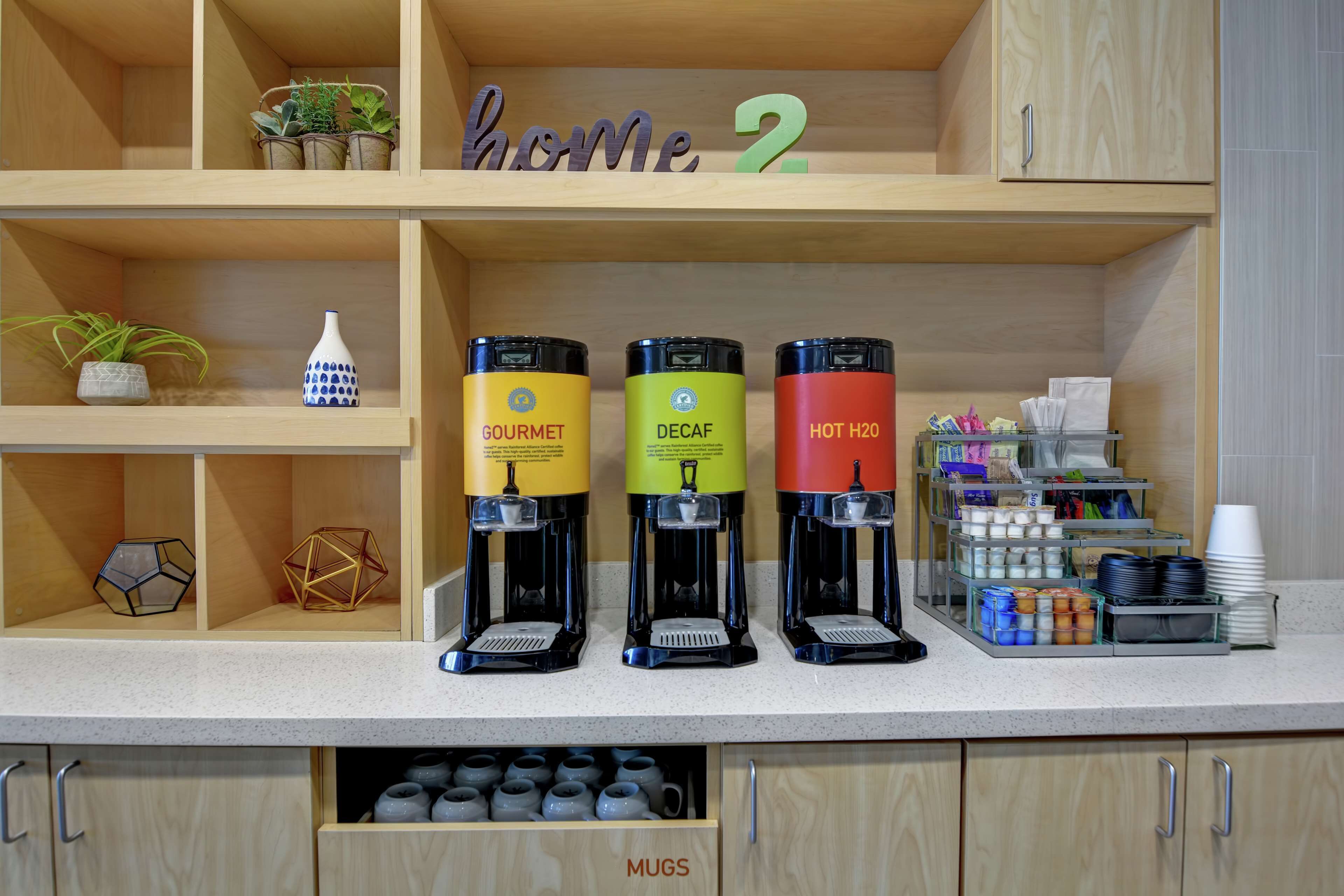 A kitchen counter with three coffee makers labeled 'GOURMET,' 'DECAF,' and 'HOT H2O.' There are shelves above with potted plants and a 'home 2' sign.