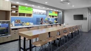 A modern kitchen area with a long wooden table and chairs, equipped with a coffee machine, refrigerator, and various food dispensers. - Home2 Suites by Hilton Denver South/Centennial Airport (Centennial)