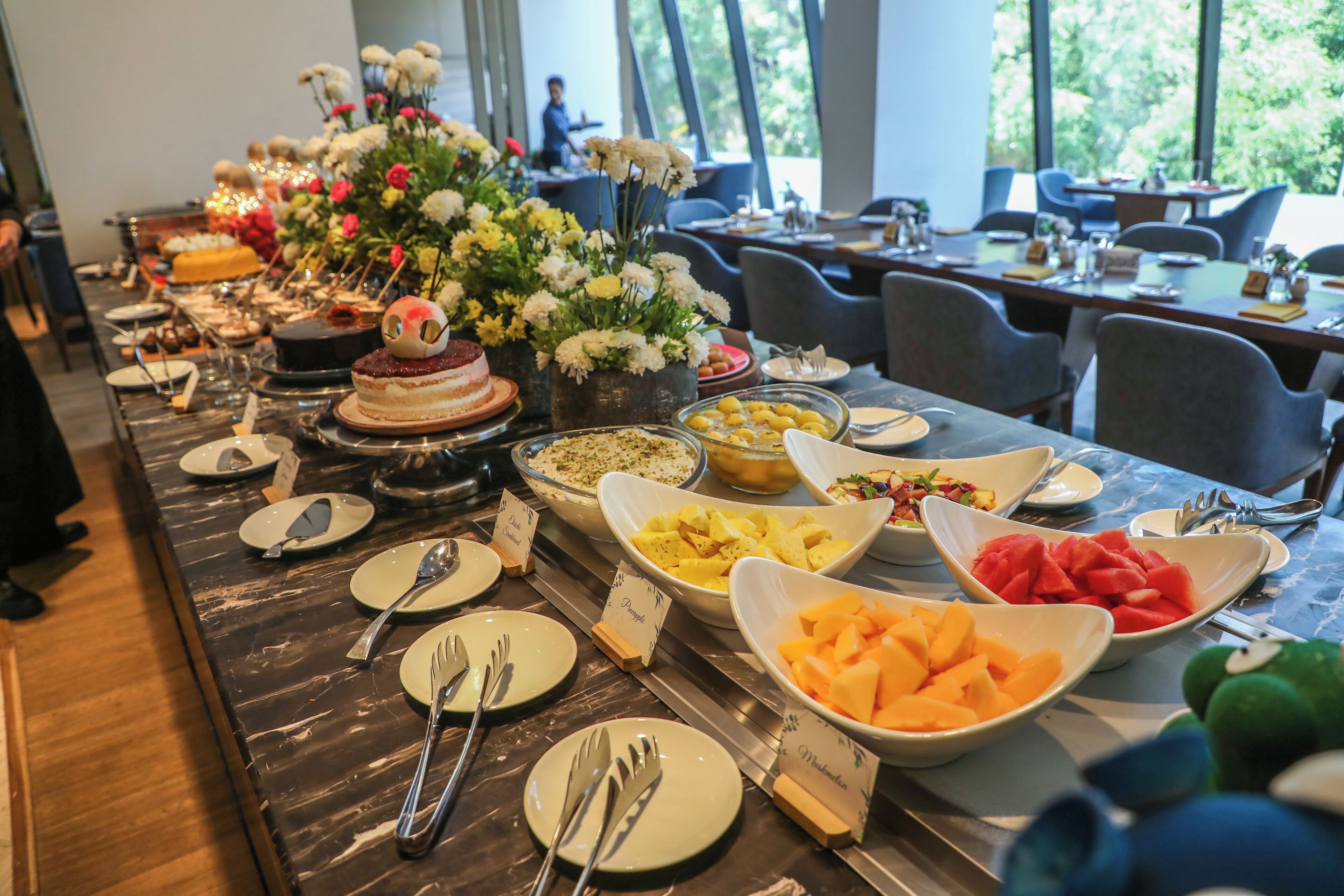 Mesa de buffet com diversos pratos e um bolo, em um salão bem iluminado com grandes janelas.