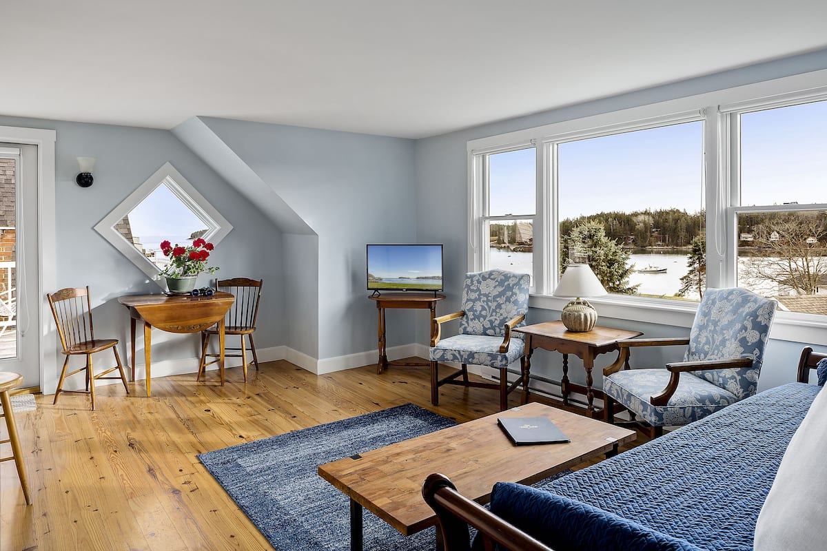 A cozy living room with a wooden floor, a blue sofa, a wooden coffee table, and a TV on a stand. There is a window with a view of a lake and trees.