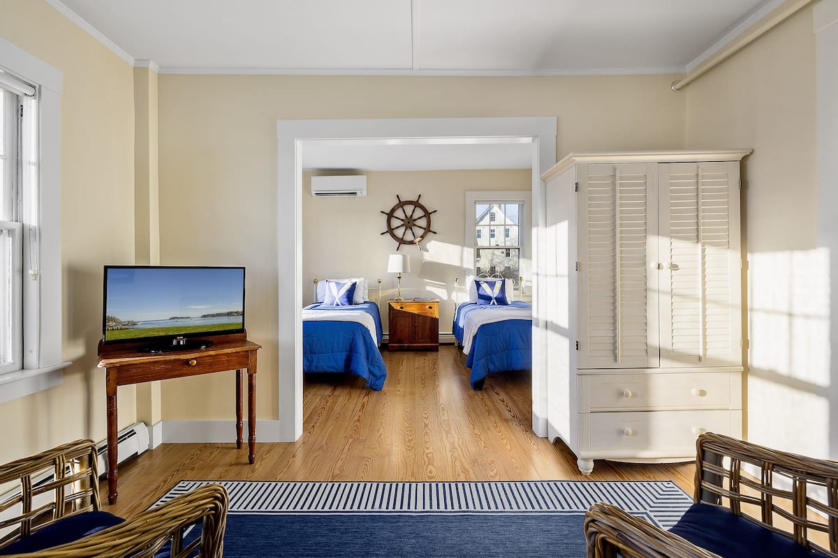A room with two beds, a television on a wooden table, a wardrobe, and a window with a view of a house.
