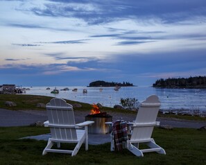 Duas cadeiras Adirondack brancas de frente para um corpo d'água, com barcos e uma ilha ao longe.