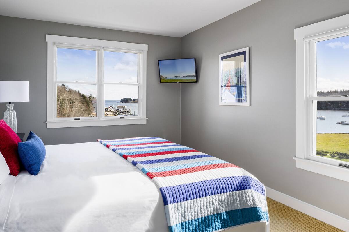 A bedroom with a bed, a TV, and two windows offering views of a body of water and trees.