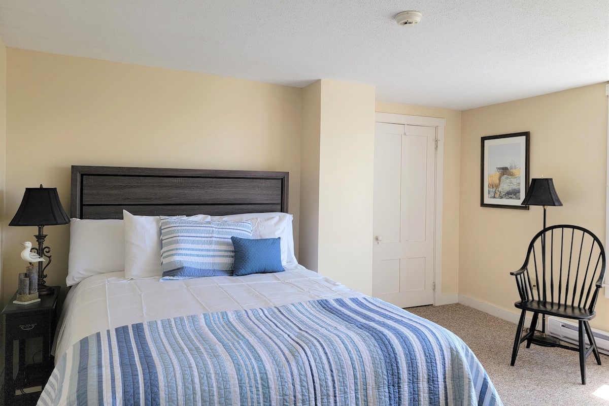 A bedroom with a bed, a nightstand, a chair, a door, and a wall-mounted picture.