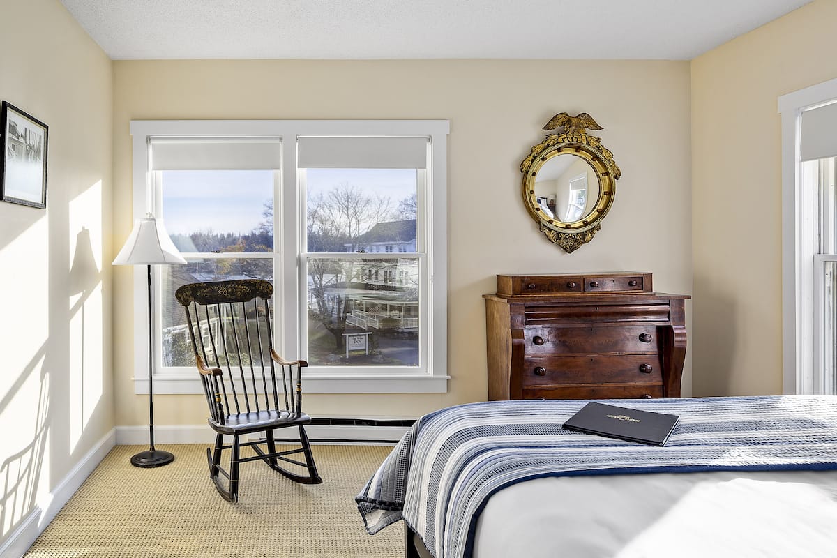 A bedroom with a large window, a rocking chair, a dresser, and a bed with a book on it.