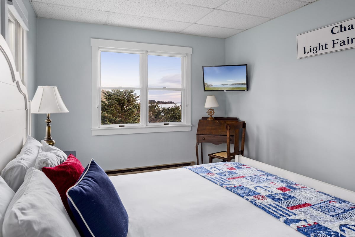 A bedroom with a bed, a desk, a chair, a TV, and a window with a view.