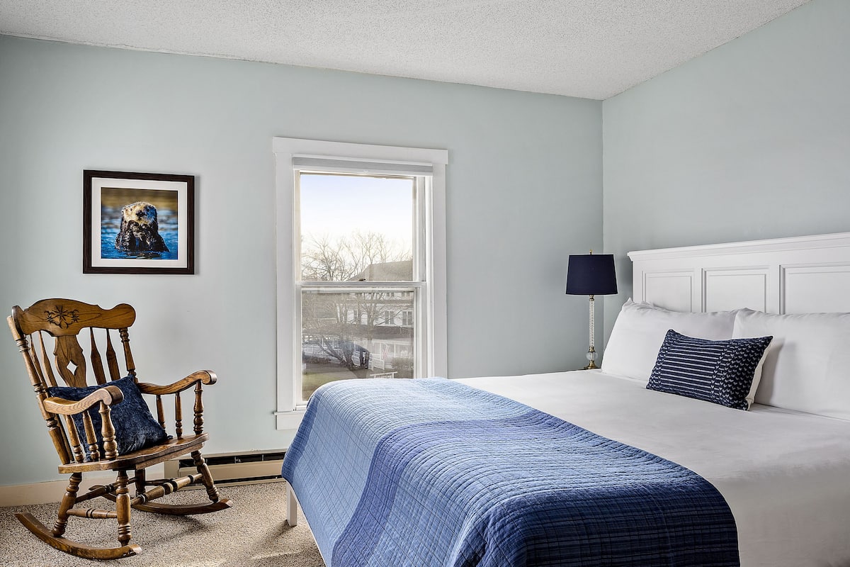 A bedroom with a bed, a rocking chair, a framed picture, and a window.