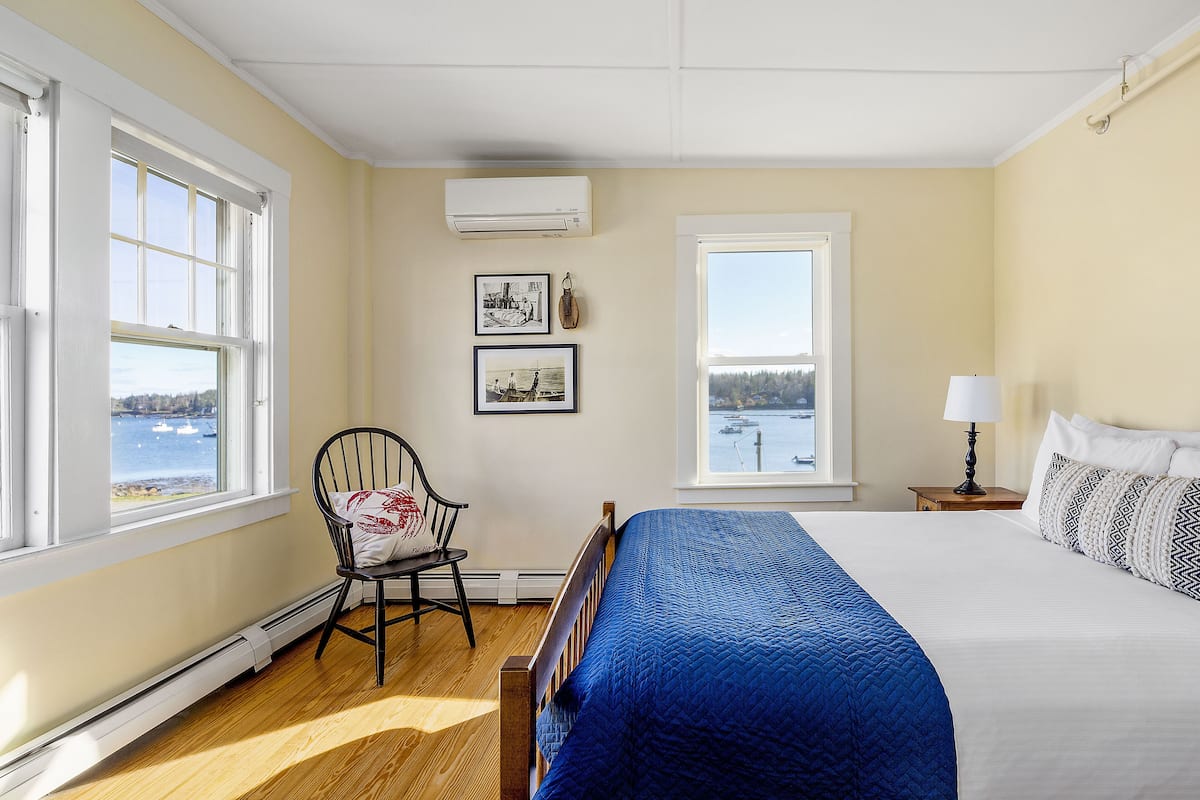 A bedroom with a large bed, a chair, a window with a view of the water, and an air conditioning unit.