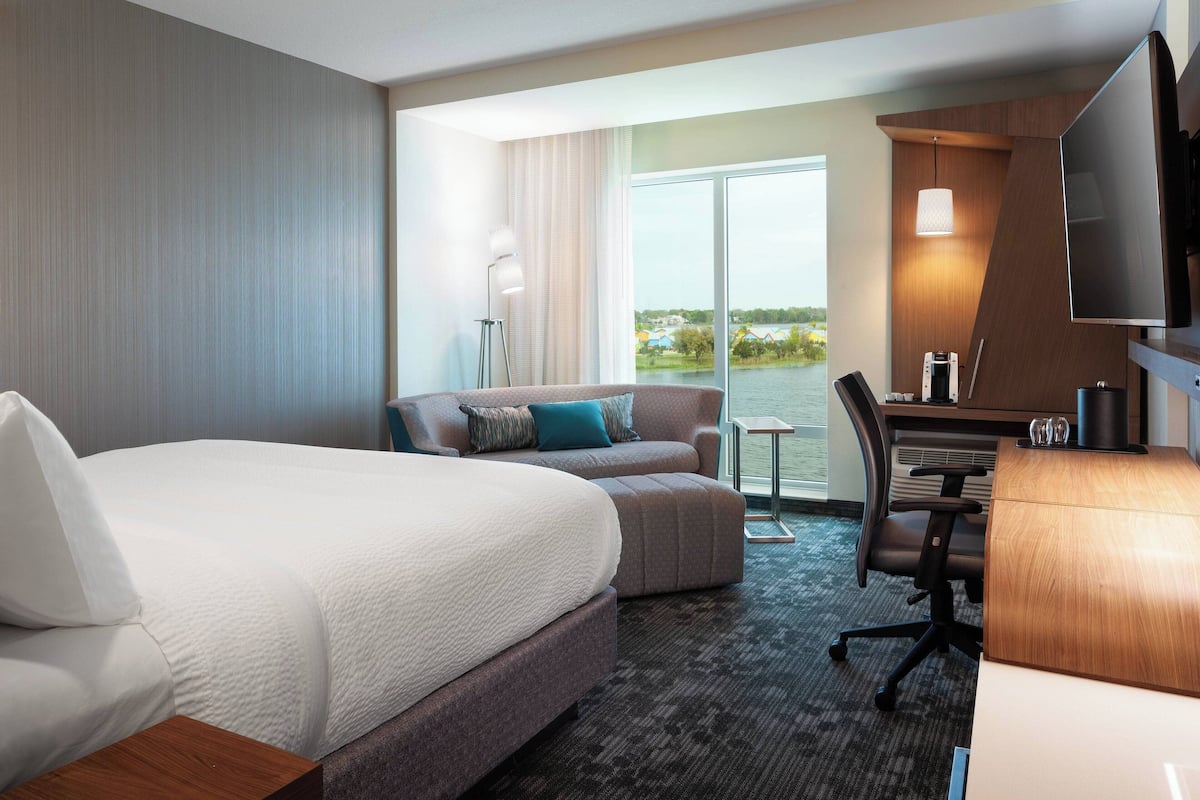 Une chambre d’hôtel avec un grand lit, un canapé, un bureau avec une chaise, une télévision et une vue sur une étendue d’eau, visible à travers une grande fenêtre.