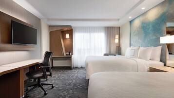 A hotel room with a desk, two beds, a flat-screen TV, a desk lamp, and a large window with curtains.