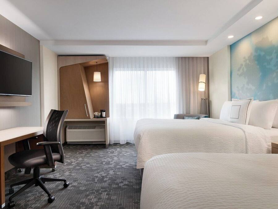 A hotel room with a desk, two beds, a flat-screen TV, a desk lamp, and a large window with curtains.