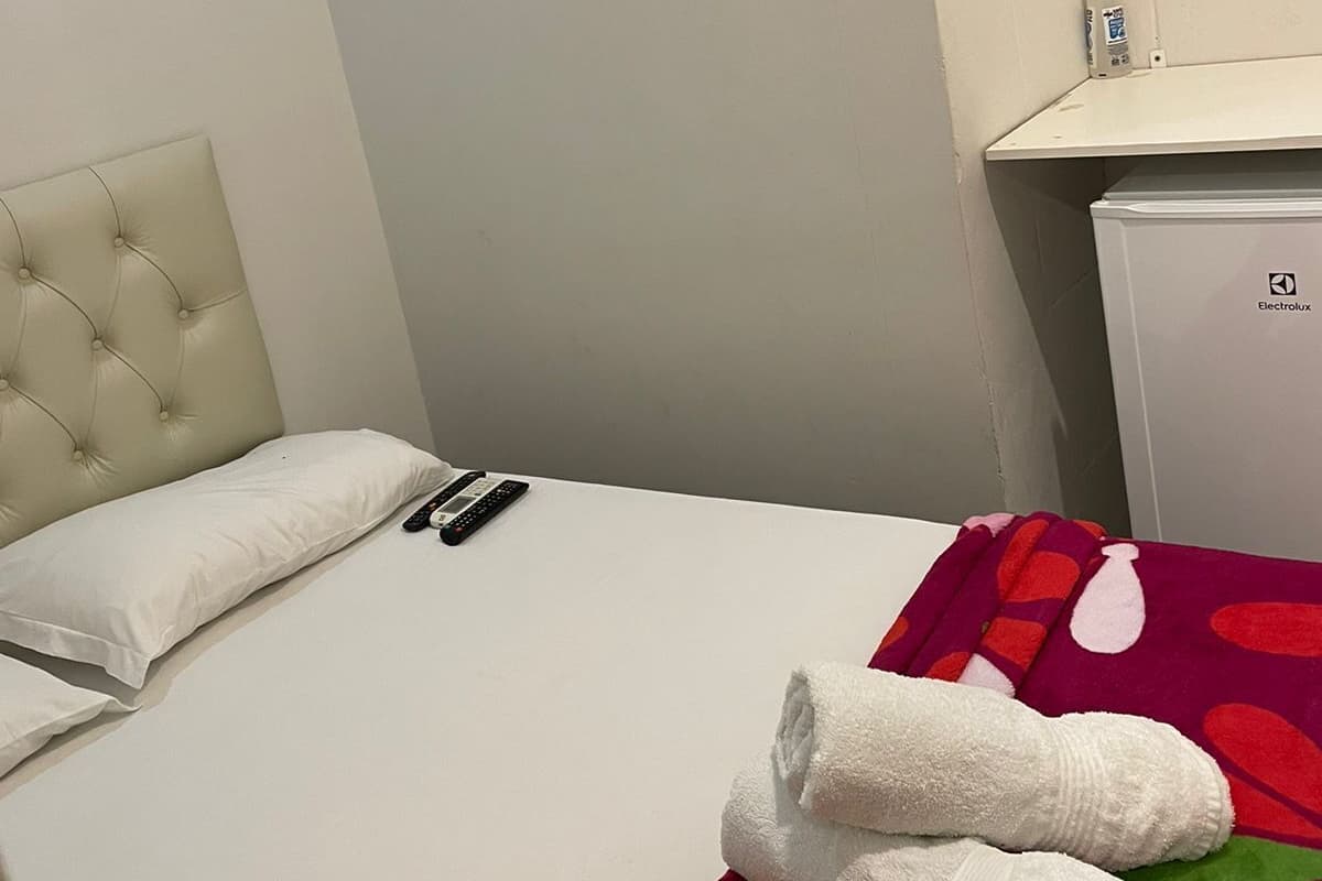 Quarto de hotel com cama, televisão na parede, mini-frigorífico e um frasco de álcool em gel no criado-mudo.