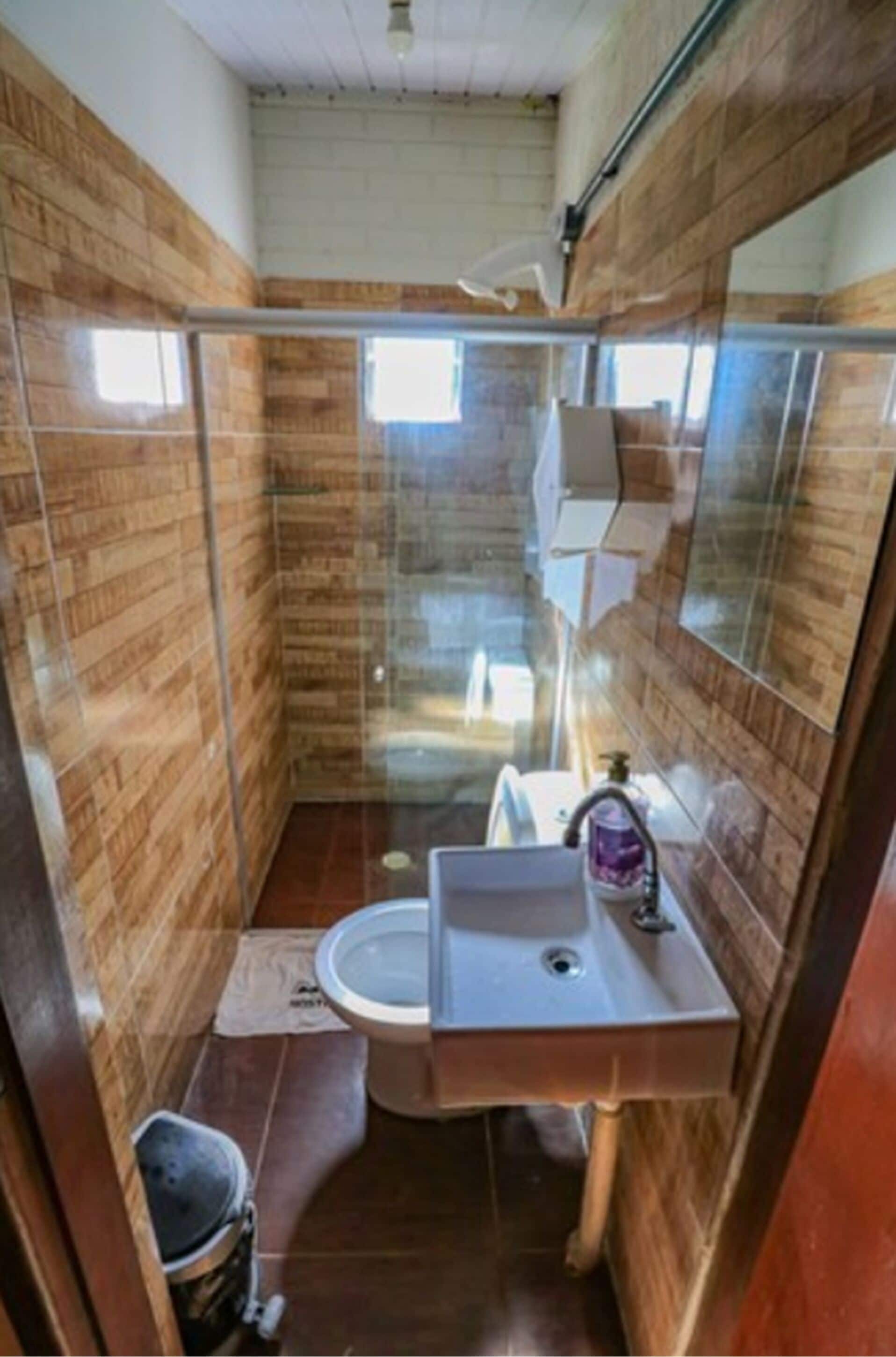 Un baño compacto con paneles de madera, mampara de ducha de vidrio, lavamanos y espejo.