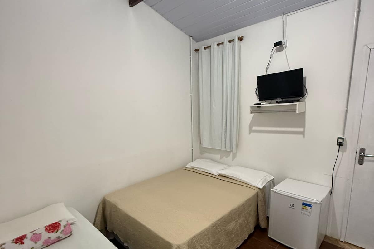 Quarto pequeno e limpo com uma cama, mesa de cabeceira, televisão presa na parede e um frigorífico pequeno.