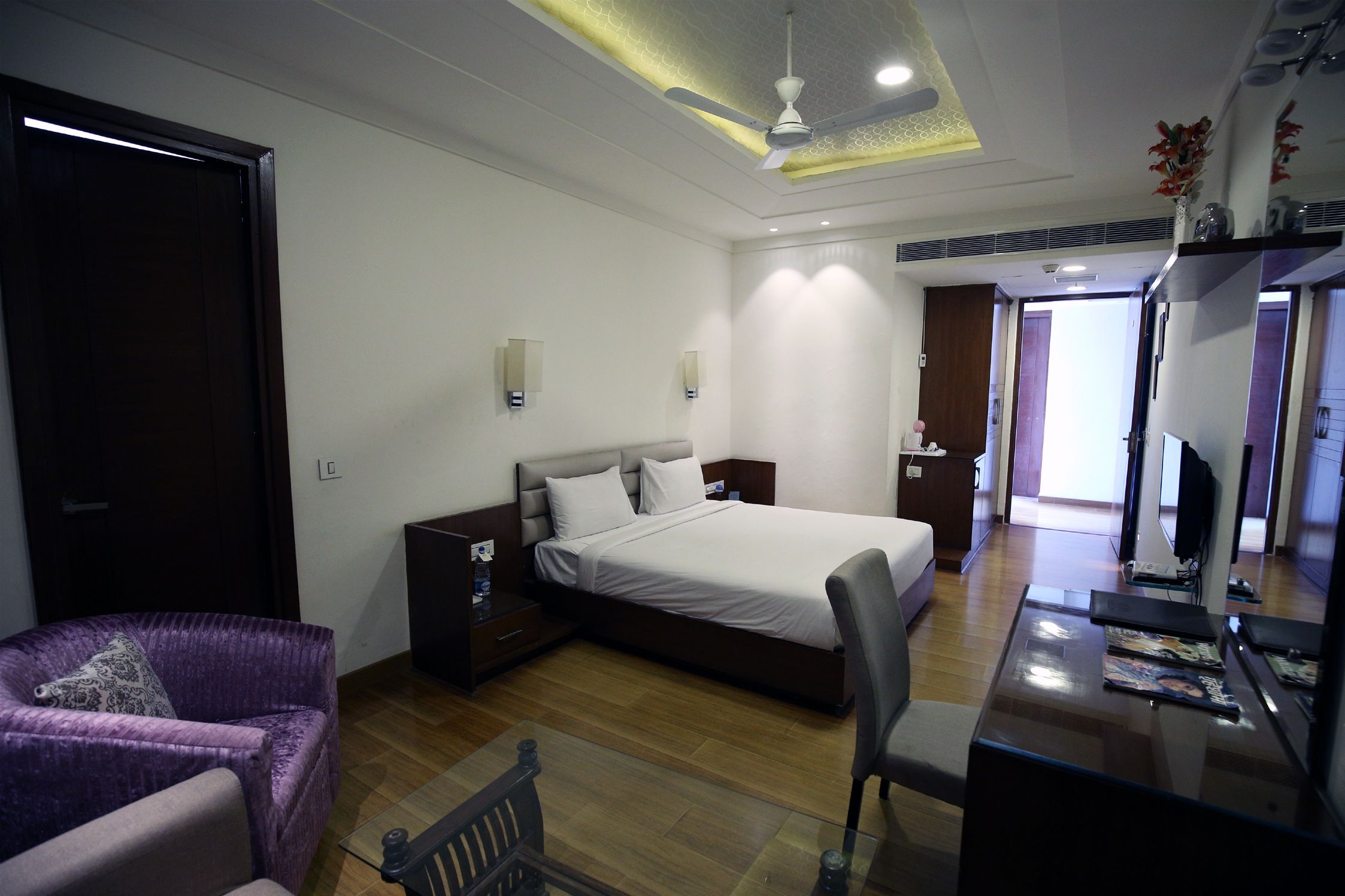 A hotel room with a bed, a desk with a chair, a purple armchair, a wooden floor, and a ceiling fan.