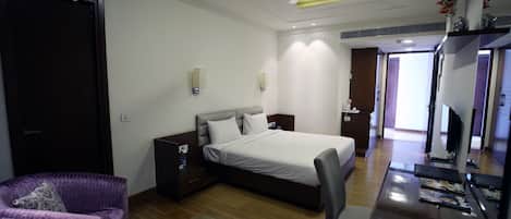 A hotel room with a bed, a desk with a chair, a purple armchair, a wooden floor, and a ceiling fan.