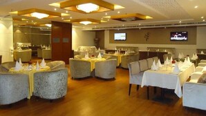 A restaurant with round tables and cushioned chairs, a television mounted on the wall, and a ceiling with recessed lighting. - Royal Inn (Muktsar)