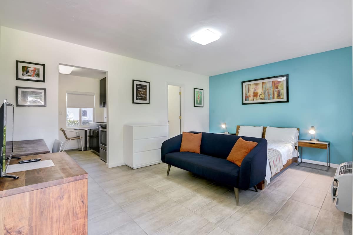 A modern room with a blue accent wall, a black sofa with orange cushions, a bed with white bedding, a desk with a computer, and a small table with a lamp.