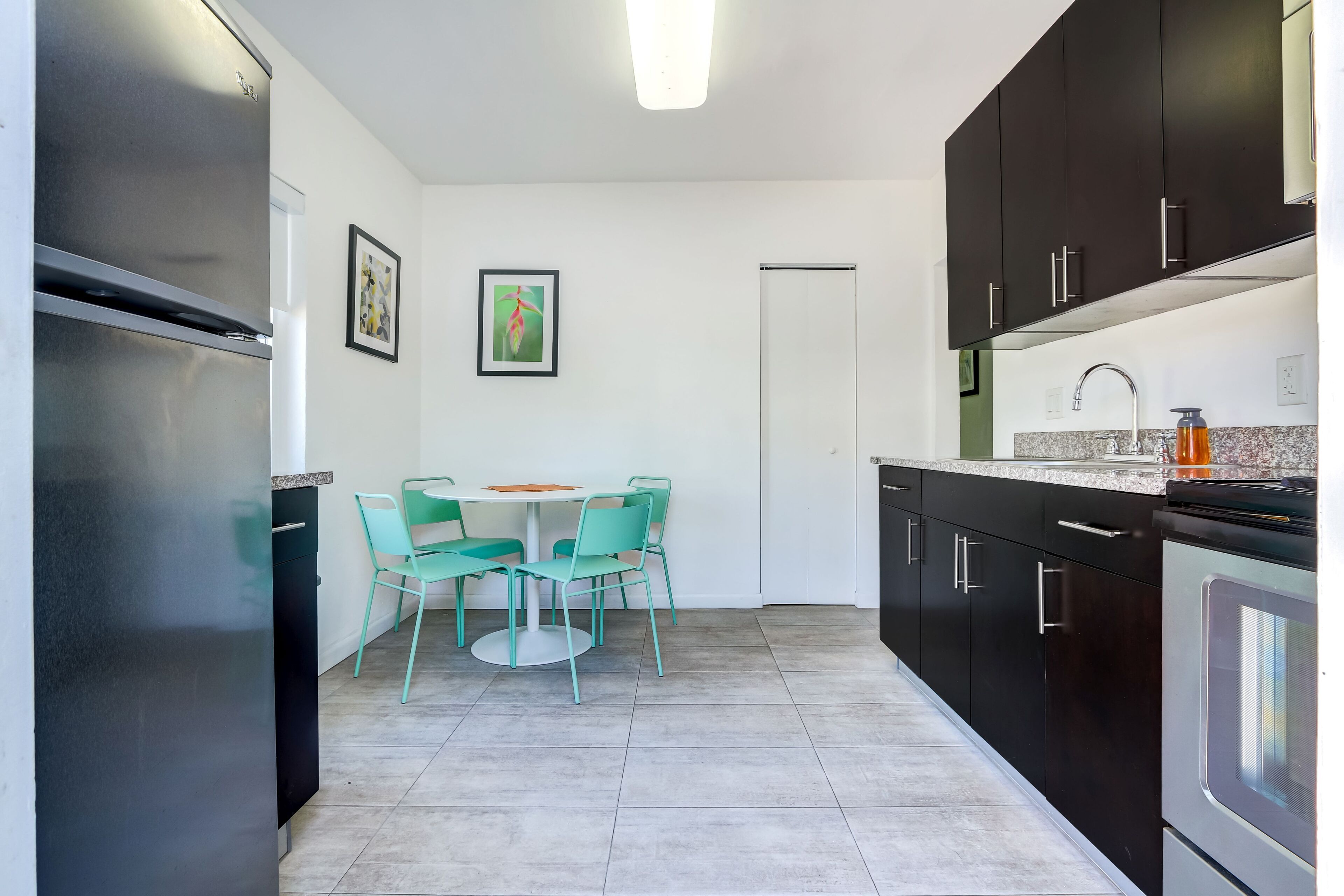 A modern kitchen with black cabinets, a stainless steel refrigerator, a dining table with four chairs, and a small window.