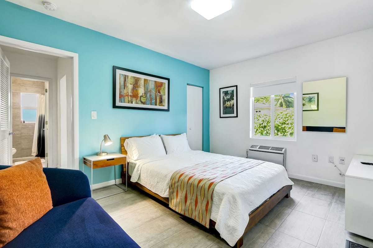 A bedroom with a bed, a desk, a chair, and a window with a view of greenery.