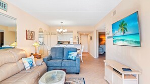 A living room with a sofa, armchair, and a TV stand. There is a kitchen in the background with a dining area.