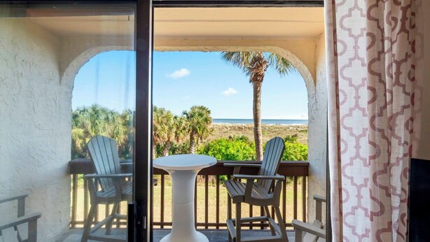 Een balkon met twee stoelen en een tafel, met uitzicht op een strand met palmbomen en de oceaan.