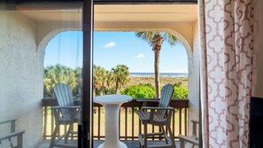 A balcony with two chairs and a table, overlooking a beach with palm trees and the ocean. - Oceanfront 2BR Getaway with Balcony, Pools, and Free Activities (St. Augustine)