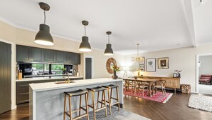 Une cuisine moderne avec des meubles de couleur sombre, un îlot central doté de tabourets, un éclairage suspendu et un coin repas agrémenté d’un tapis rouge.