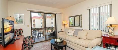 Un salon avec un canapé, une table basse et un téléviseur posé sur un meuble. Il y a un balcon d’où l’on peut admirer la vue sur les immebles, et il est doté d’une rambarde.