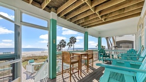 Una terraza techada con sillas y una mesa de madera, con vista a una playa con palmeras y el océano.