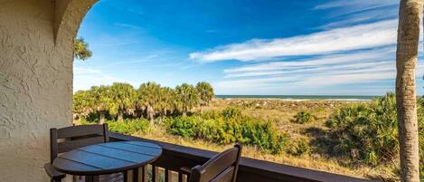 A wooden deck with a table and chairs overlooking a beach with palm trees and ocean.