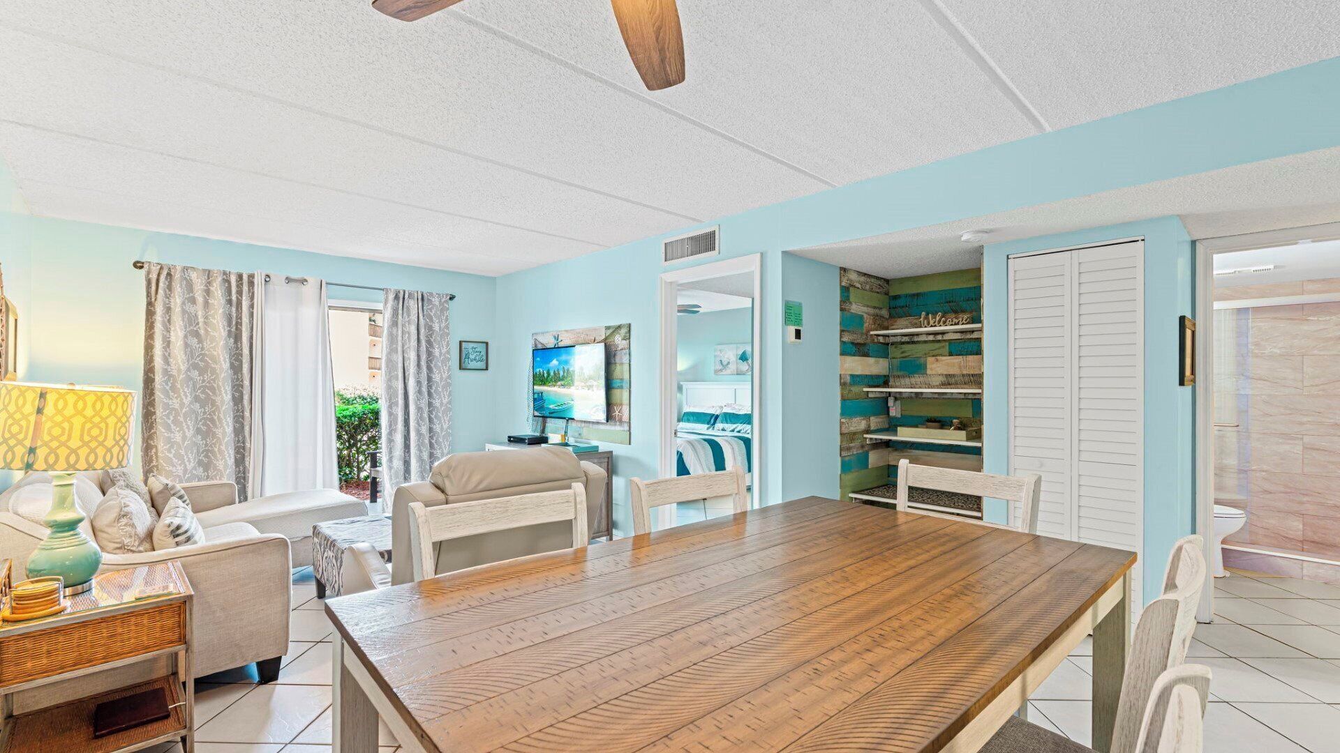 A spacious living room with a wooden dining table, a sofa, and a TV. There is a window with curtains and a view of greenery outside.
